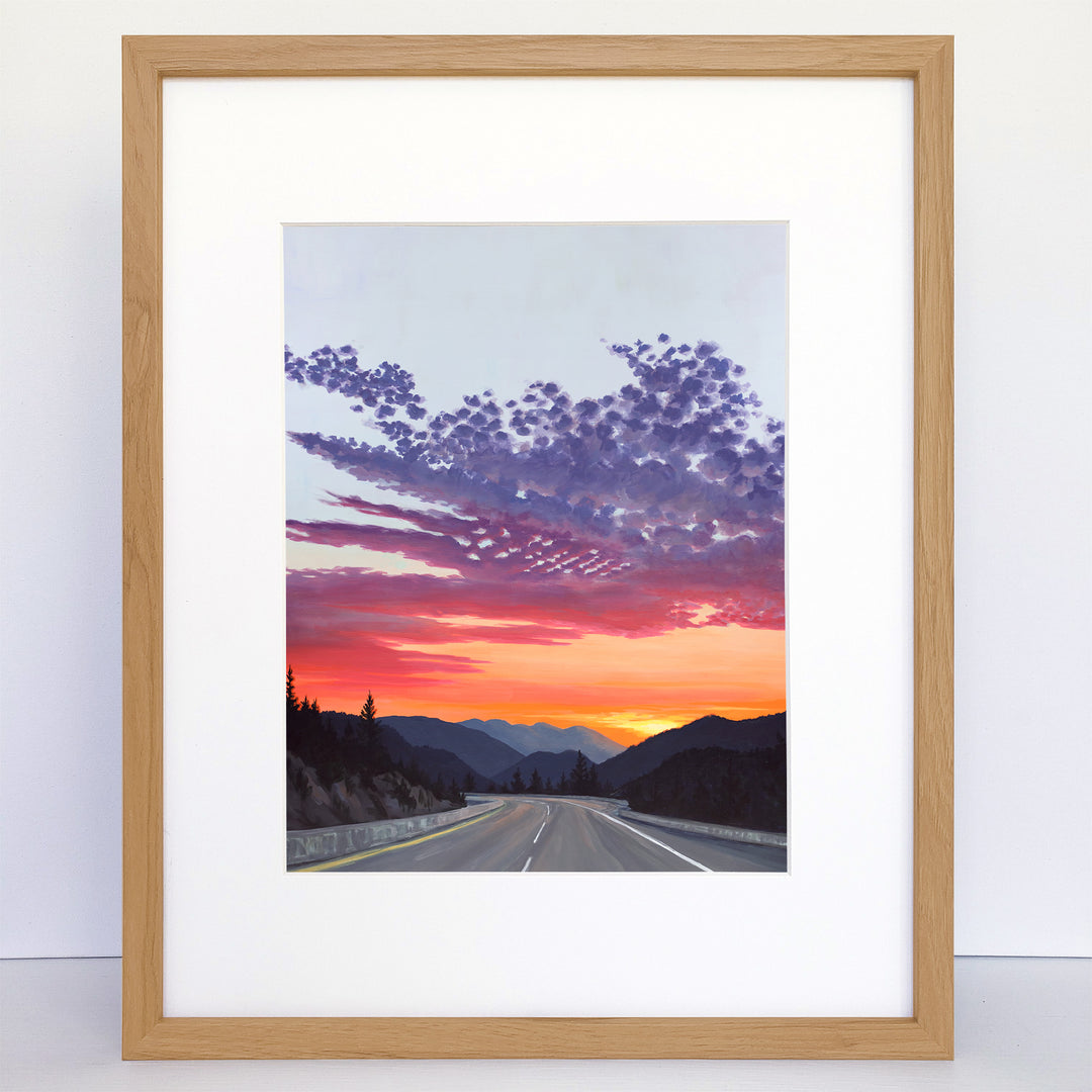 Framed art print of a sunset over a mountain highway. Bright orange, pink, yellow and purple.