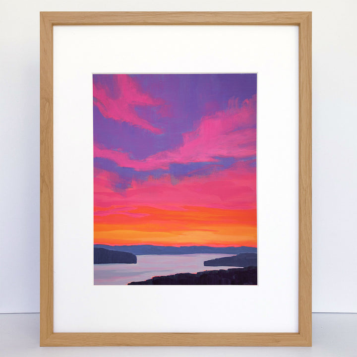 A framed vertical print showing a vibrant pink, orange, and purple sunset over a river. 