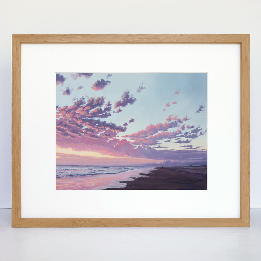 A framed art print showing an ocean beach with a pink and purple sunset. 