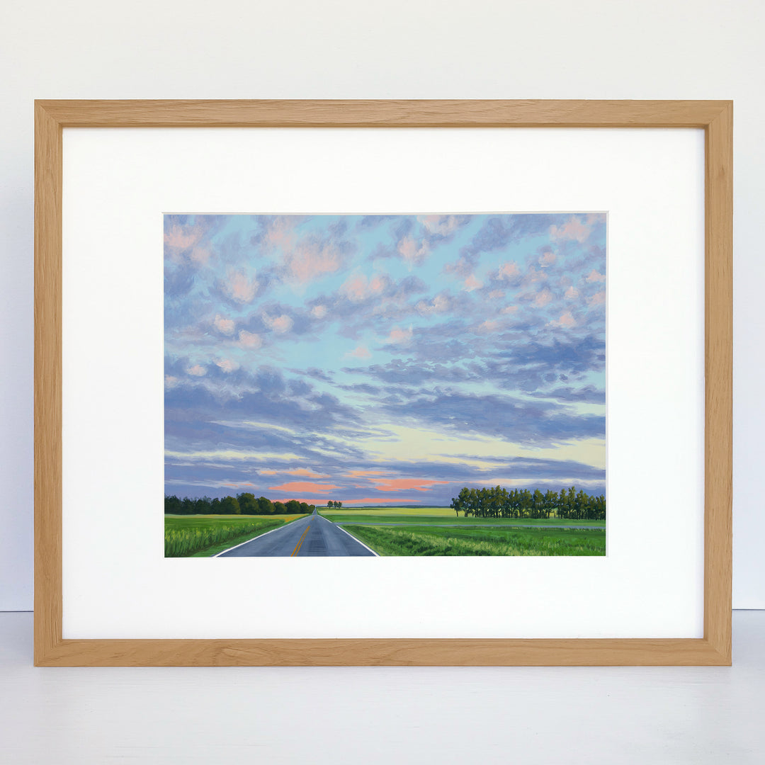 A framed art print showing a country scene with green fields, trees on the horizon and a pink and purple pastel sunset.