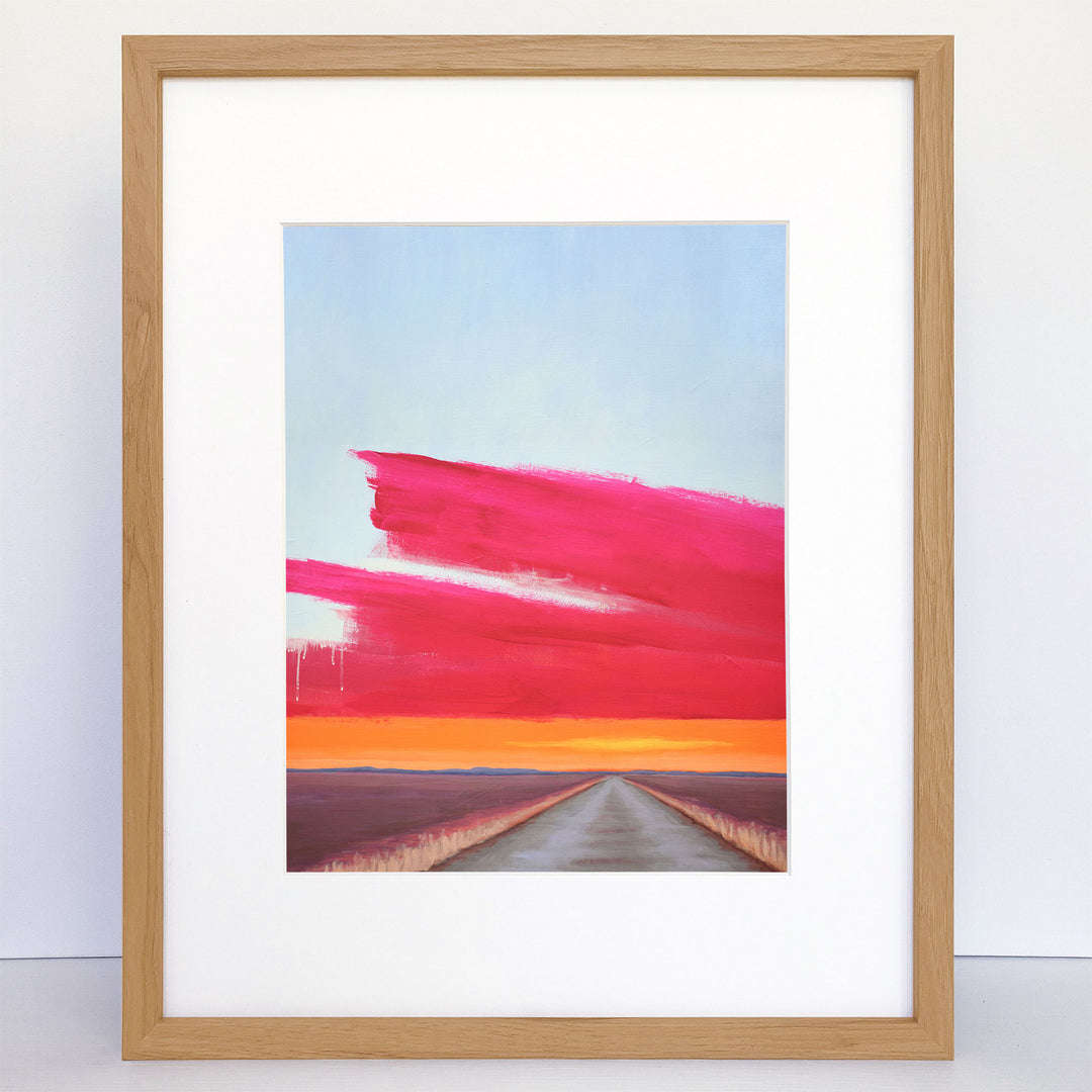 A framed art print showing a dirt road and a bright orange and pink sunset.