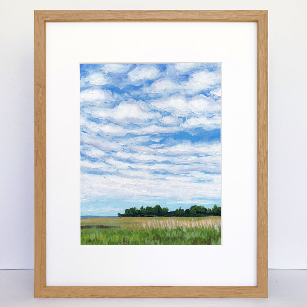 Framed art print of green field and trees with blue sky and white, puffy clouds.