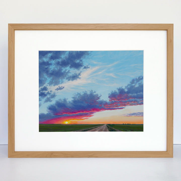 A framed horizontal painting showing a vibrant sunset over green fields and a dirt road.