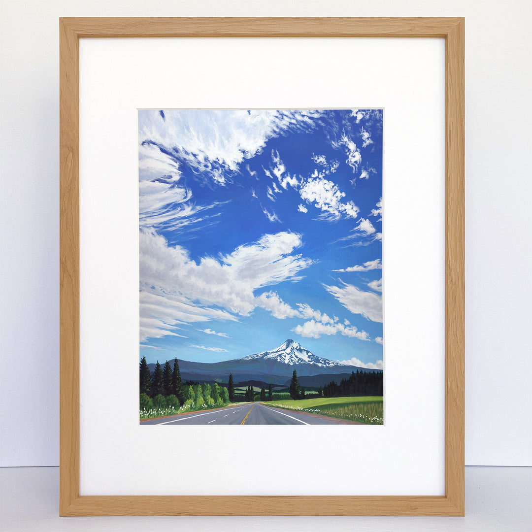 Frame vertical art print of a summer day, country road and a big mountain. The sky is blue with lots of white clouds. Fields and trees in the foreground.