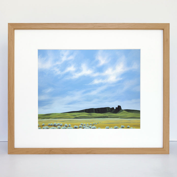 Framed horizontal art print showing a green field, a rock formation on the horizon, and a blue sky with wispy clouds.
