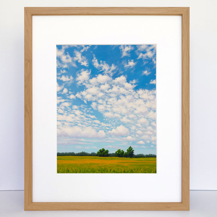 A framed vertical art print of a summer day with blue sky and puffy clouds. A field of grass and yellow flowers. 
