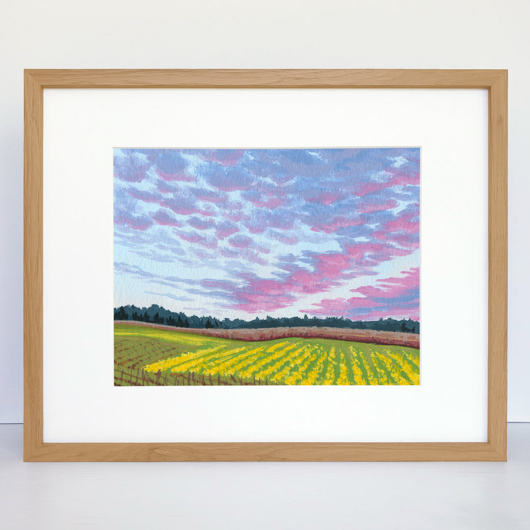 A horizontal framed art print of a pink and purple sunset and yellow crops. 