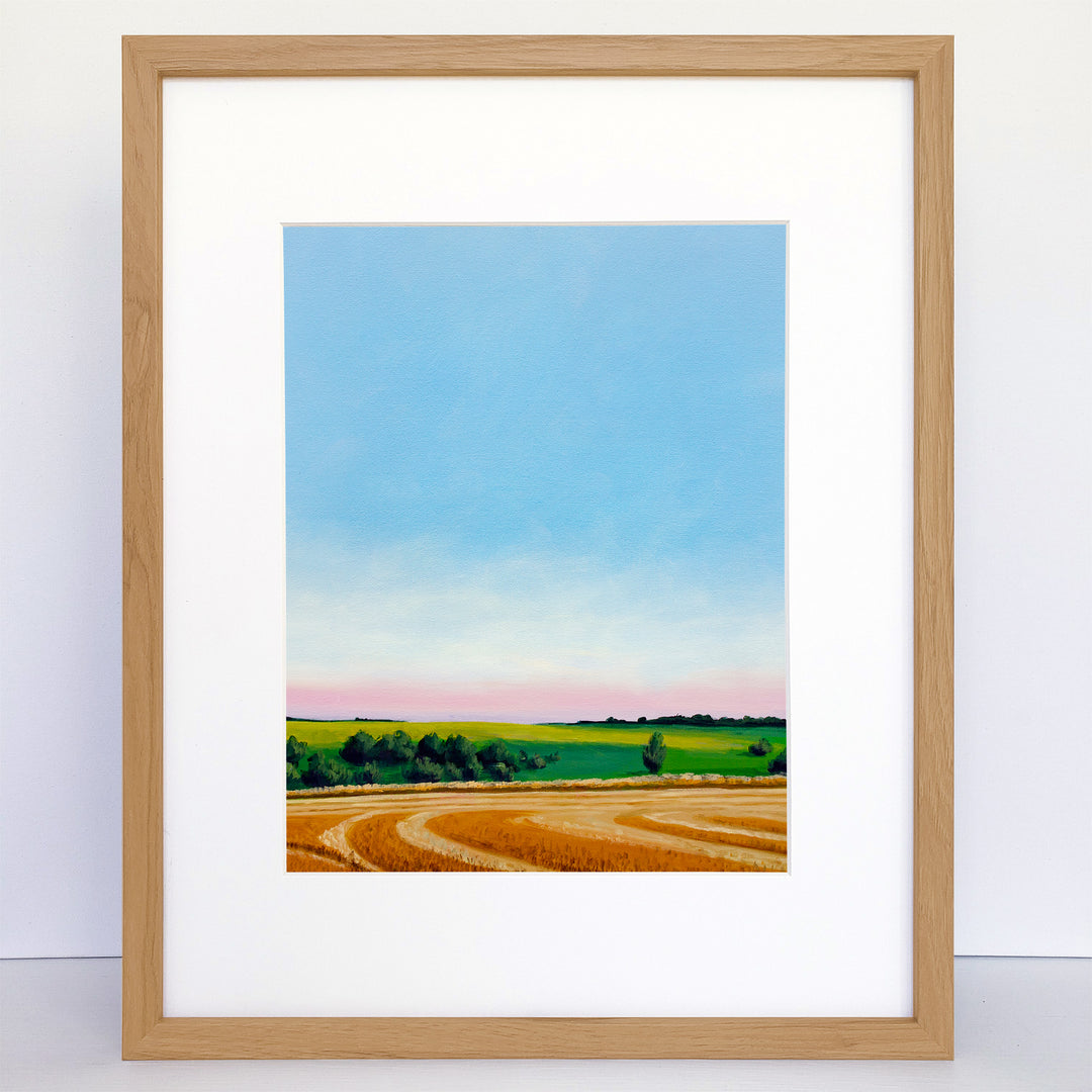 Framed landscape art of wheat field, green hill and pink and blue sky.