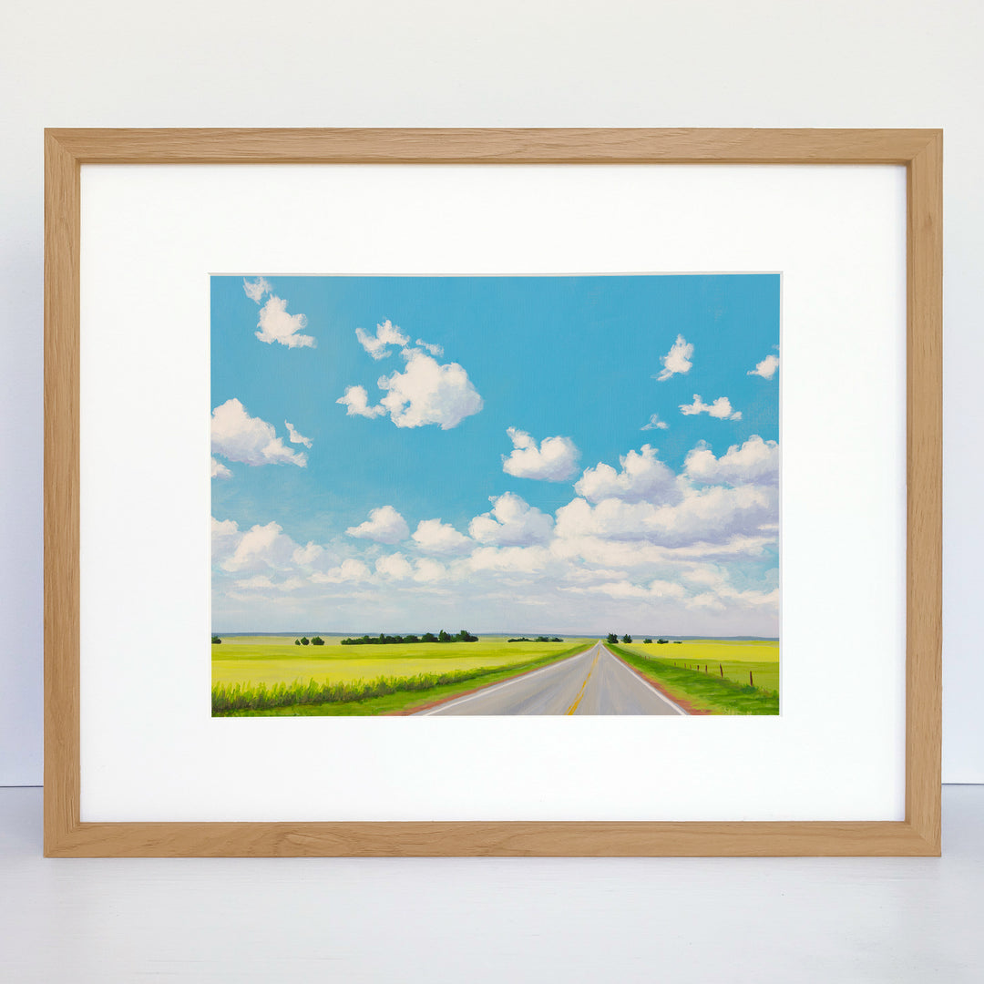 A framed art print showing a two-lane road with green fields on both sides, a blue sky, and white puffy clouds.