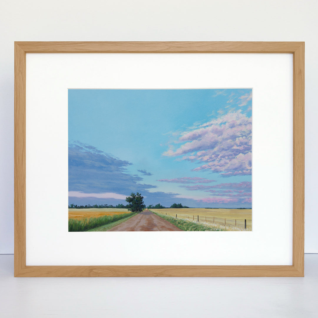 A horizontal framed print showing a dirt road, end-of-summer fields and a pastel sunset.